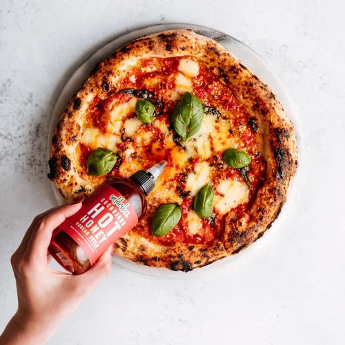 Person drizzling WilderBee hot honey from a bottle onto a pizza with basil leaves