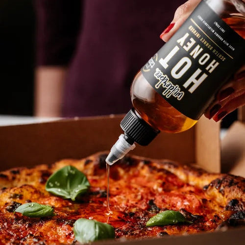 Person drizzling WilderBee hot honey from a bottle onto a pizza with basil leaves.