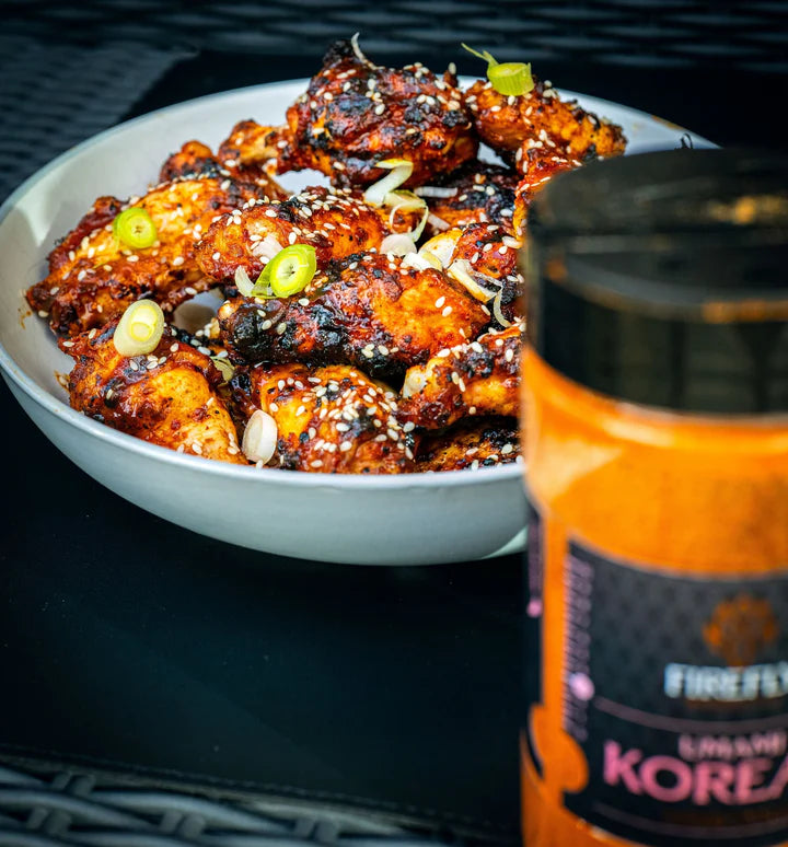 Bowl of grilled chicken wings with a jar of Firefly Korean BBQ rub on a dark surface.