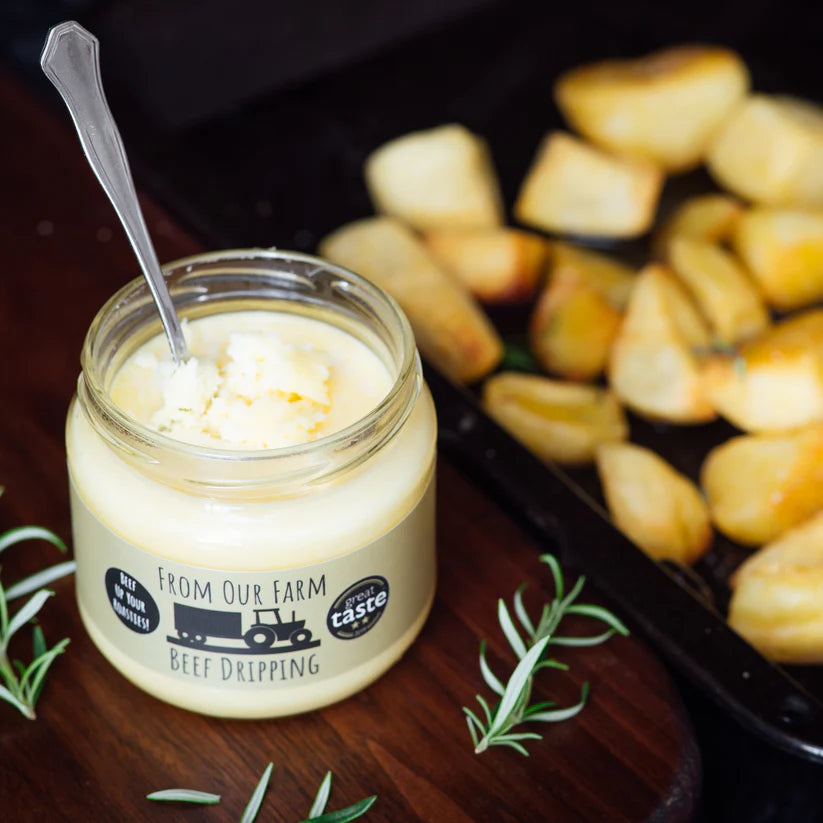 Jar of open beef dripping on a wooden board with rosemary next to a tray of roast potatoes