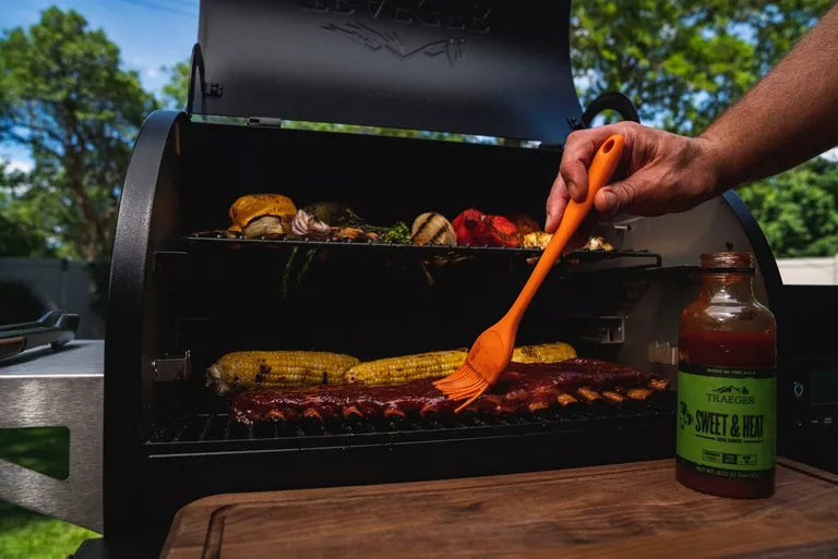 Person basting grilled vegetables and meats with a bottle of sauce on a wooden cutting board.