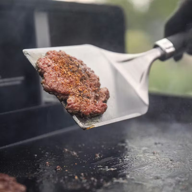 Hamburger patty being flipped on a grill with a Weber stainless spatula.
