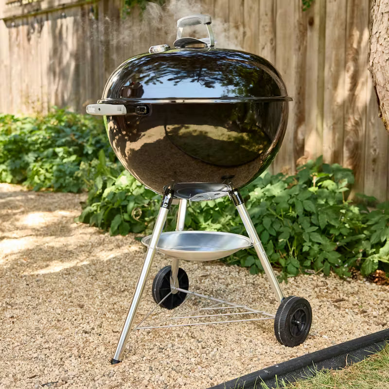 Black Weber kettle grill on wheels in a backyard setting with plants and a wooden fence.